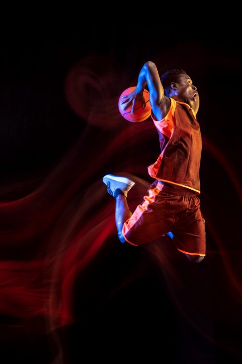 Mysterious nature. African-american young basketball player of red team in action and neon lights over dark studio background. Concept of sport, movement, energy and dynamic, healthy lifestyle.
